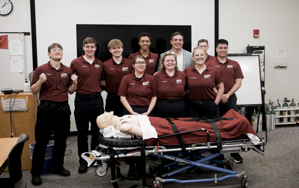 Full EKU EMC paramedic cohort group photo with Dr. Andrew Napier, simulation mannequin, and Ferno stretcher in the training lab
